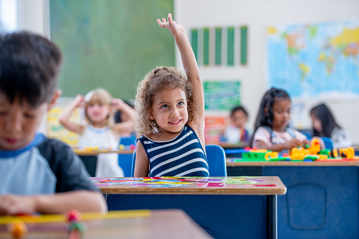 A young elementary student sits at her desk with her hand raised as she waits to ask a question.  She is dressed casually and seated among her peers.