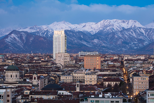 The city of Turin, with its palaces churches skyscrapers, on a windy winter evening. It is the blue hour, wind clouds sweep over the distant mountain ridges, while city lights illuminate Turin's buildings, squares and monuments.