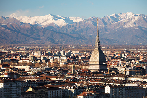 The city of Turin, dominated by its symbol, the Mole Antonelliana, on a windy winter afternoon. In the mountains there is little snow and the wind clouds sweep over the distant mountain ridges, while the city hustles and bustles amid its buildings, squares and monuments.