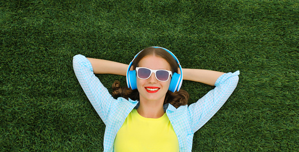 Portrait of happy smiling young woman listening to music in headphones while lying on grass in summer park