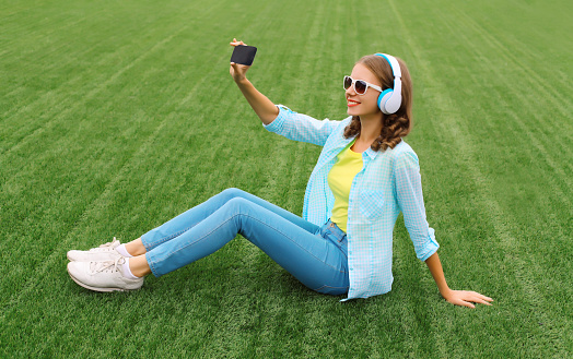 Happy young woman taking selfie with smartphone listening to music in headphones while lying on grass in summer park