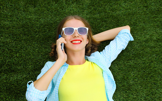 Portrait of happy smiling young woman calling on mobile phone while lying on green grass in summer park, top view