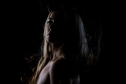Woman with long hair standing in dimly lit environment. Beautiful blond girl side lit silhouette portrait.
