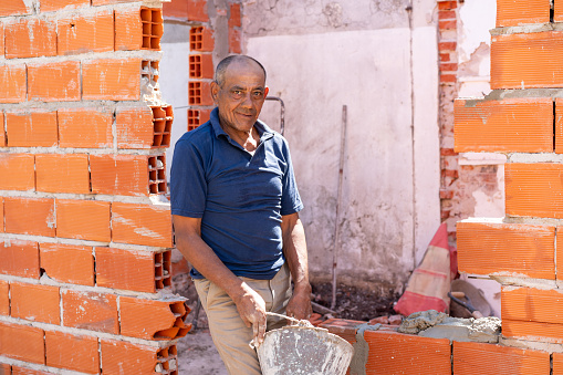 Mature construction worker building a brick wall and holding a bucket with cement while facing the camera slightly smiling - Construction industry concepts