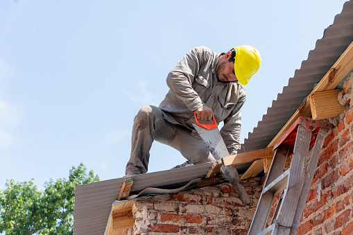 Construction worker using a hand saw to cut a piece of wood while installing roof tiles - Construction industry concepts