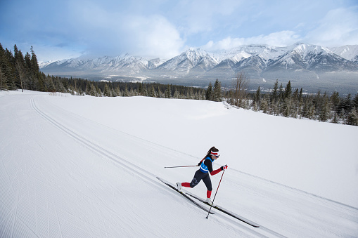 A young woman competes in a cross-country ski race at the Canmore Nordic Centre Provincial Park in Alberta, Canada. She is doing the classic-style technique, wears bib number 14, and has a maple leaf on her race suit. She wears a headband, sports sunglasses, and uses classic-style skis and boots.