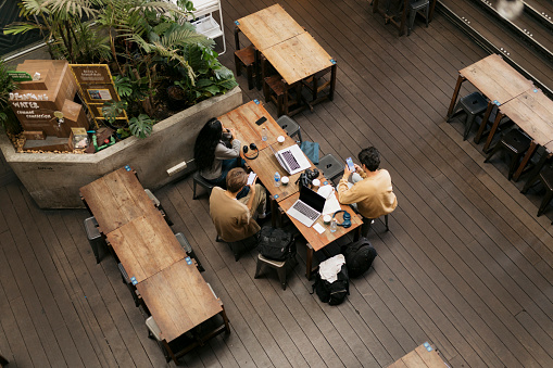Top view of young people sitting around coffee table using their mobile phones. View from above of college students sitting at cafe table with laptops, headphones, camera and disposable coffee cups.