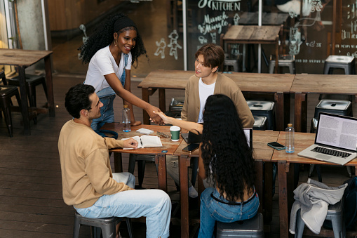Young african woman shaking hands with female at cafe table with friends sitting by. Woman meeting and greeting friends sitting at coffee shop.