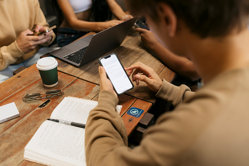 Over the shoulder view of young male student using mobile phone while sitting at cafe table with his friends. Cropped shot of young people using their mobile phones sitting at coffee shop with book and laptop.