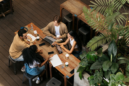 High angle view of group of young students gather around a cafe table, chatting and working on an assignment. They look focused and engaged, exchanging ideas and notes in a relaxed cafe atmosphere.