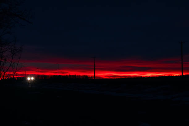Early morning in prairies. Dramatic red and blue sunrise in the rural area and the highway with power poles along it. and lonely vehicle with lights.