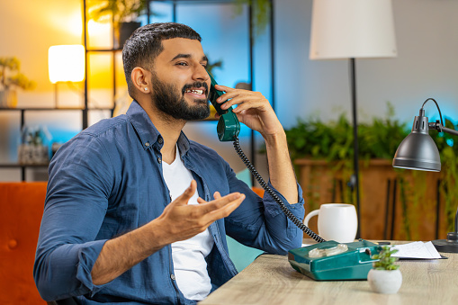 Bearded Indian man freelancer working, talking with client on retro old-fashioned wired telephone at home office workplace table. Salesman in casual clothes. Remote distant job. Employment occupation.