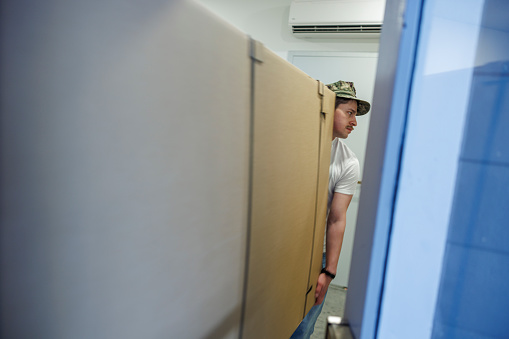 Furniture moving: Hispanic man in a cap moves a heavy box with furniture and takes it to an elevator. Personal point of view