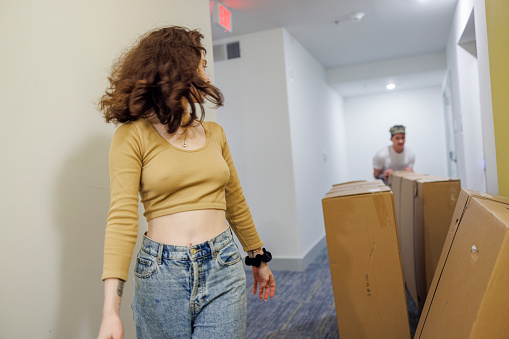 During the move in, a young Caucasian brunette woman is showing the way to a man mover with cardboard boxes, from the lift to the apartment