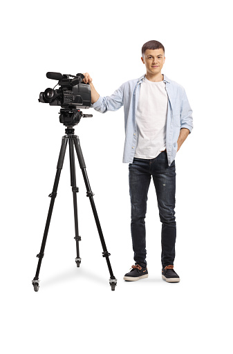 Full length portrait of a young camera operator with a camera on a tripod stand isolated on white background