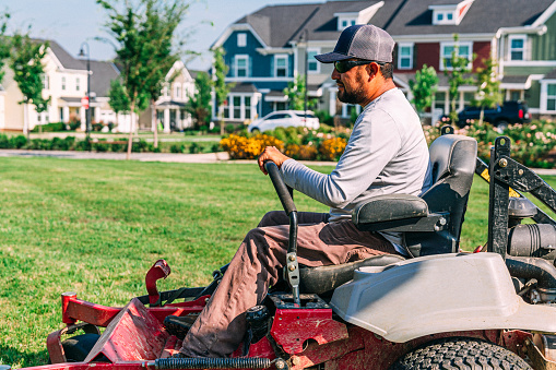 Hispanic Man Driving a Professional Riding Lawnmower for a Landscaping Company at an Apartment Complex or Public Park on a Sunny Morning in the Summer, USA
