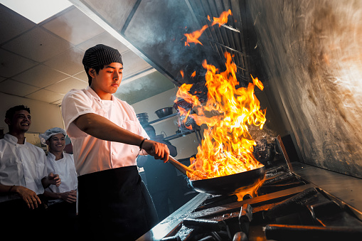 Young Asian chef in the restaurant kitchen making flambe in the frying pan.