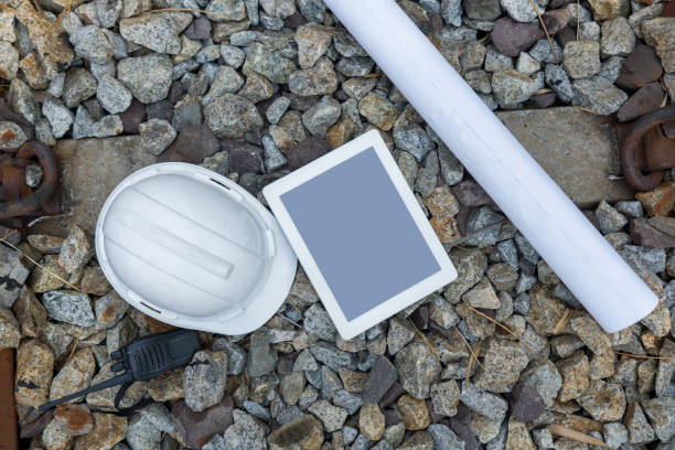 A tablet computer, hardhat, walkie-talkie and train route chart lie on ballast stone in railroad next to railroad sleeper and rail beam. Top view