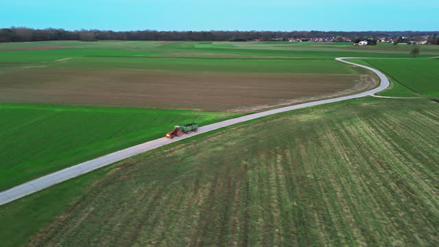 Aerial Drone Footage of Tractor with Trailer Moving on Road amidst Green Agricultural Landscape against Blue Sky