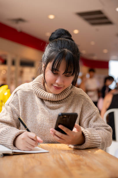 istock A woman is multitasking, taking notes in her book while reading some information on her phone. 2044422220