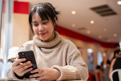 A beautiful, positive Asian woman in a comfy sweater is chatting with someone on her smartphone while sitting in a cafe. people and wireless technology concepts