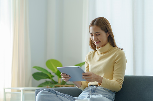 Bright and cute Asian woman using tablet to listen to music and other entertainment Relaxing at home happily watching movie and playing game at home.