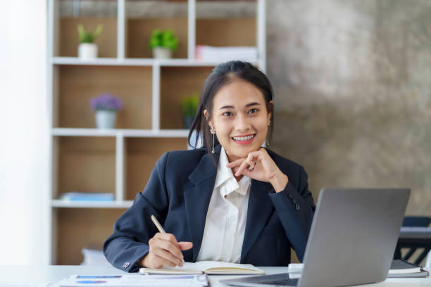 istock Smiling beautiful young Asian businesswoman sitting with laptop and computer while doing some paperwork at the office. Looking at camera. 2044488303