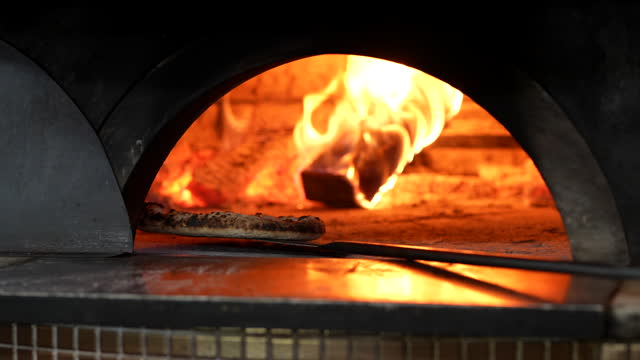 In the cropped shot, a chef is seen adjusting the pizza paddle inside a brick oven fire.