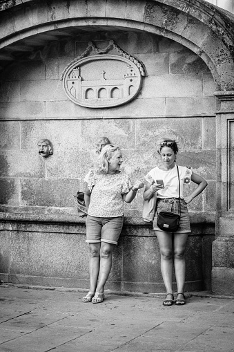 Pontevedra, Spain - August 4, 2022: Two women chat sitting in one of the city's granite stone fountains, with their coat of arms in relief.
