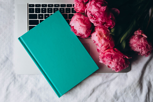 Laptop, green book and bouquet of pink peonies flowers on a white bed, top view.
