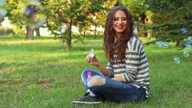 Young Woman Blowing Bubbles While Sitting On The Grass In The Park