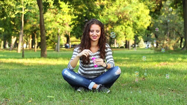 Young Woman Blowing Bubbles While Sitting On The Grass In The Park