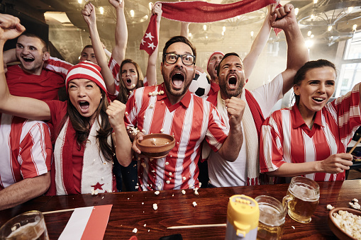 Large group of cheerful fans celebrating the goal of their favorite sports team while watching a game on TV in a bar.