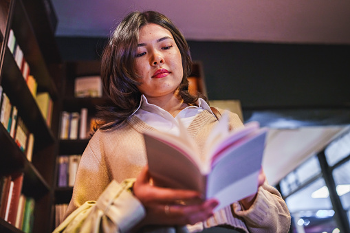 Young woman reading a book in front of a bookshelf in bookstore.