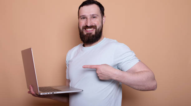 istock Excited happy bearded man pointing finger at blank screen laptop computer isolated over beige background. 2044797185