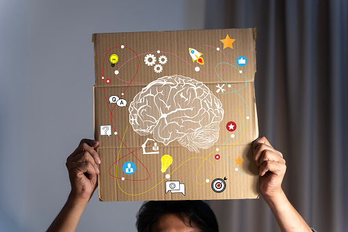 A businessman holds a piece of cardboard above his head and on the cardboard there is a drawing of a brain representing thoughts. Imagination at work. Sit and work in the office until dark.