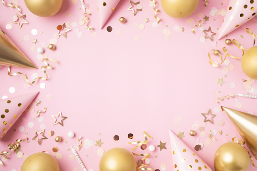 Birthday party festive background with golden decoration from balloons, carnival hat, confetti on pastel pink table top view.