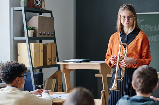 Young female music teacher holding trumpet standing in classroom in front of kids conducting class