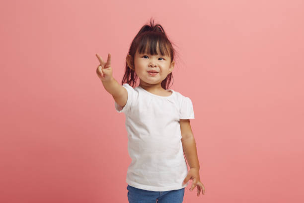 linda ittle menina asiática sorri e mostra o gesto v em pé sobre o fundo isolado rosa. - 6184 - fotografias e filmes do acervo