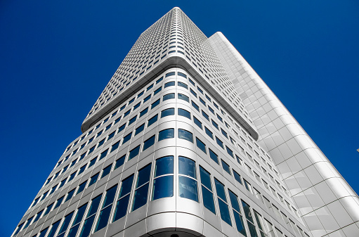 White-silvery shiny high-rise facade of a modern office tower in sunlight and with a blue cloudless sky in oblique bottom view