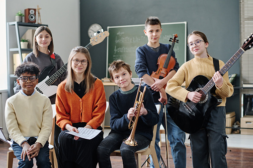Group of boys and girls taking part in school orchestra and their young music teacher posing for camera with instruments in classroom
