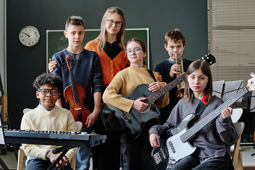 Group portrait of modern middle school orchestra members and music teacher posing for camera with instruments in their classroom