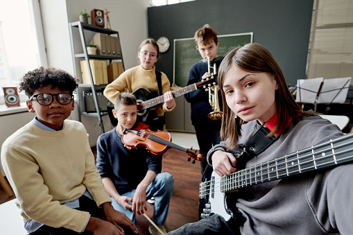 Modern teen girl with bass guitar holding camera taking selfie with her friends at music class