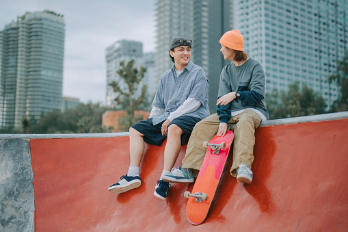 Asian Chinese skateboard couple holding skateboard sitting side by side at skateboard park resting  on wall feeling relax