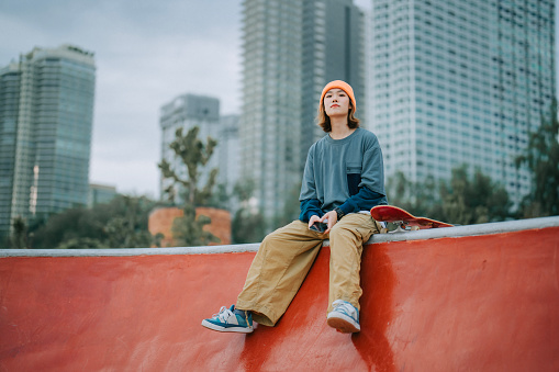 Asian Chinese female skateboarder sitting at skateboard park looking away