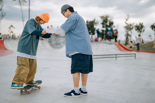 Asian Chinese boyfriend holding girlfriend hands practising ollie skateboarding in skateboard park weekend morning