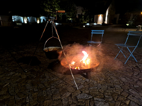 night grilling and roasting on an open fire. the metal bowl is filled with wood and is portable. paved area with blue chairs. meat is cooked in a cauldron suspended over the fire