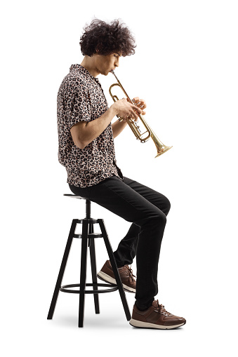Full length profile shot of a young male artist sitting on a chair and playing a trumpet isolated on white background