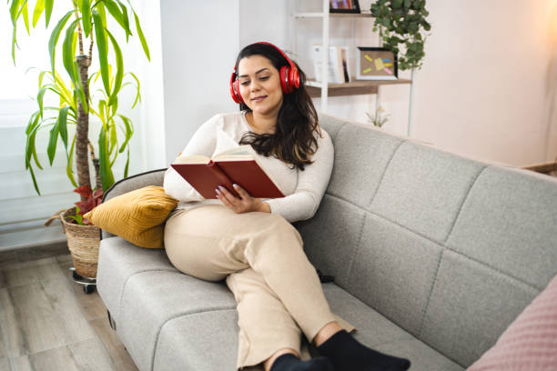 istock Woman with wireless headphones reading a book on a sofa 2045073415