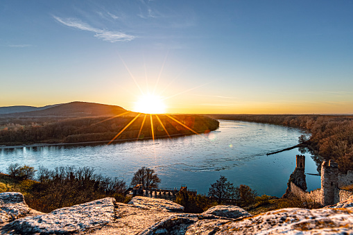 Bratislava, Slovakia - December 6, 2018: As the sun sets over the horizon, casting a golden glow across the landscape, the Danube River from the vantage point of Devin Castle is a breathtaking sight to behold.\n\nThe river reflects the vibrant hues of the sky, painting the water with shades of orange, pink, and purple. Silhouettes of boats drift lazily along the tranquil surface, while the distant hills are bathed in the soft light of dusk.